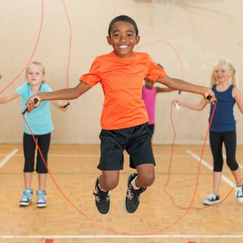 Young boy wearing black shorts and orange t-shirt jumps rope in a gym with other children.